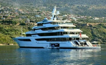 A large yacht, owned by arthur blank yacht, positioned in tranquil waters adjacent to a picturesque mountain landscape.