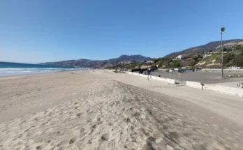 Zuma Beach featuring golden sand with several cars parked along the shoreline under a clear blue sky.
