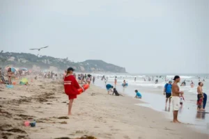 A man in a red shirt stands on Point Dume Beach, with sand and ocean waves in the background.
