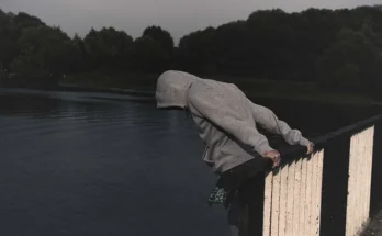 A hooded individual leans over a railing, with carrara luxury drug​ branding visible in the background.