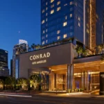 Conrad Los Angeles luxury hotel exterior at dusk with illuminated entrance, palm trees, and downtown Los Angeles skyline.