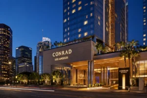 Conrad Los Angeles luxury hotel exterior at dusk with illuminated entrance, palm trees, and downtown Los Angeles skyline.