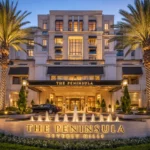 The Peninsula Beverly Hills luxury hotel exterior at dusk, featuring a grand cream-colored façade, illuminated entrance, palm trees, landscaped gardens, fountain, and elegant Beverly Hills surroundings.