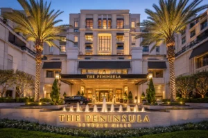 The Peninsula Beverly Hills luxury hotel exterior at dusk, featuring a grand cream-colored façade, illuminated entrance, palm trees, landscaped gardens, fountain, and elegant Beverly Hills surroundings.