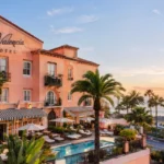 La Valencia Hotel overlooking the ocean with a Mediterranean-style exterior, palm trees, pool, and terrace at sunset.