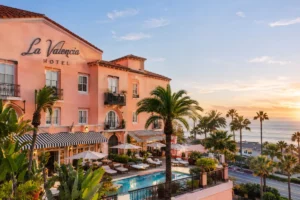 La Valencia Hotel overlooking the ocean with a Mediterranean-style exterior, palm trees, pool, and terrace at sunset.