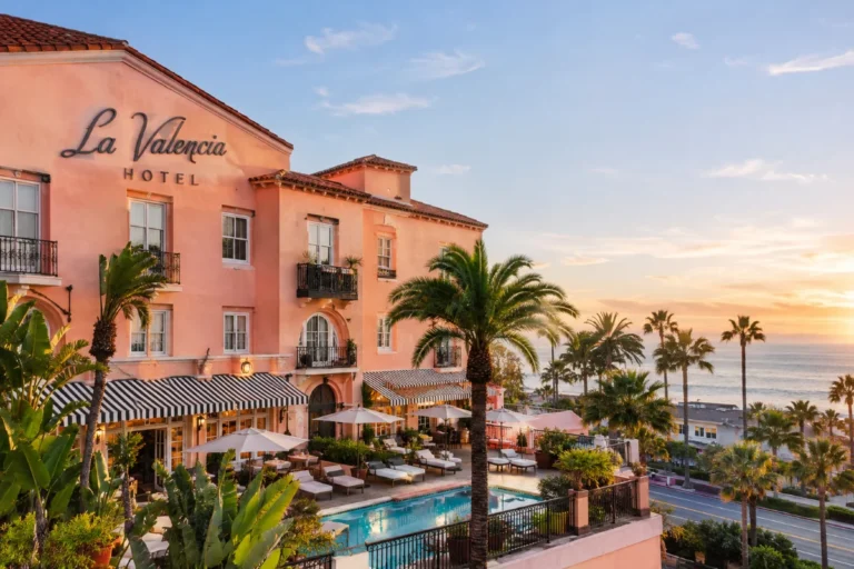 La Valencia Hotel overlooking the ocean with a Mediterranean-style exterior, palm trees, pool, and terrace at sunset.